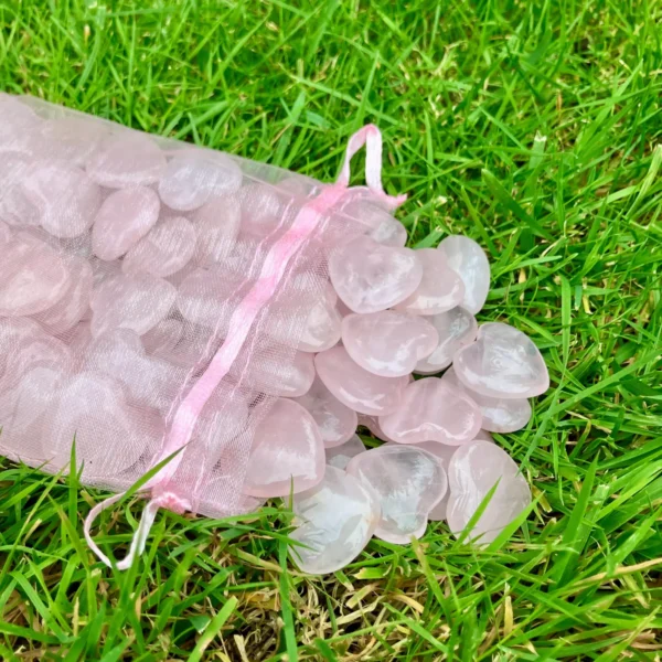 May include: A pink mesh bag filled with heart-shaped rose quartz stones. The stones are a translucent pink color and are scattered on a bed of green grass. The May include: A pink mesh bag filled with heart-shaped rose quartz stones. The stones are a translucent pink color and are scattered on a bed of green grass. The bag has a pink ribbon drawstring.