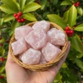 May include: A woven basket filled with raw, light pink rose quartz crystals. The crystals have a rough, natural texture and varying sizes. The basket is held in a hand, with green foliage and red berries in the background.