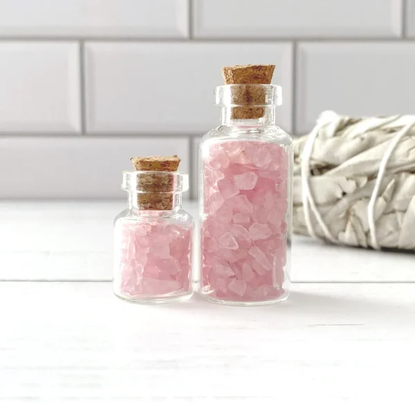 May include: Two glass bottles with cork stoppers filled with pink rose quartz crystals. The bottles are on a white wooden surface. May include: Two glass bottles with cork stoppers filled with pink rose quartz crystals. The bottles are on a white wooden surface.