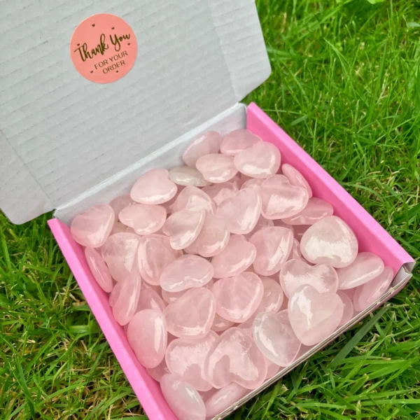 May include: A box filled with polished rose quartz heart-shaped stones. The box has a pink interior and a white exterior. A round sticker with the words "Thank May include: A box filled with polished rose quartz heart-shaped stones. The box has a pink interior and a white exterior. A round sticker with the words "Thank You For Your Order" is on the box. The stones are a soft pink color.