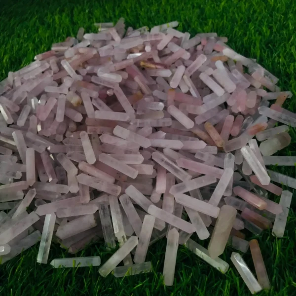 May include: A large pile of pink and clear crystal points, also known as wands, on a green grassy surface. May include: A large pile of pink and clear crystal points, also known as wands, on a green grassy surface.
