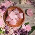 May include: A wooden bowl filled with raw, pink rose quartz crystals. The crystals vary in size and shape, with a translucent appearance. The bowl is surrounded by pink and purple dried flowers, creating a natural, earthy aesthetic.
