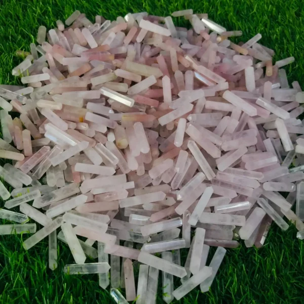 May include: A large pile of pink and clear crystal points, also known as wands, are arranged on a bed of green grass. The points are various sizes and shapes, May include: A large pile of pink and clear crystal points, also known as wands, are arranged on a bed of green grass. The points are various sizes and shapes, with some having a slight iridescent sheen.