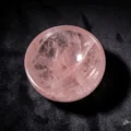 May include: A polished, round rose quartz bowl. The bowl is a soft pink color with internal veining and a smooth, reflective surface. The bowl is set against a dark background.