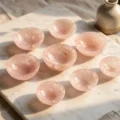 May include: A collection of eight small, round bowls made of translucent rose quartz. The bowls are a soft pink color with natural veining and variations in tone. They are arranged on a white marble surface.