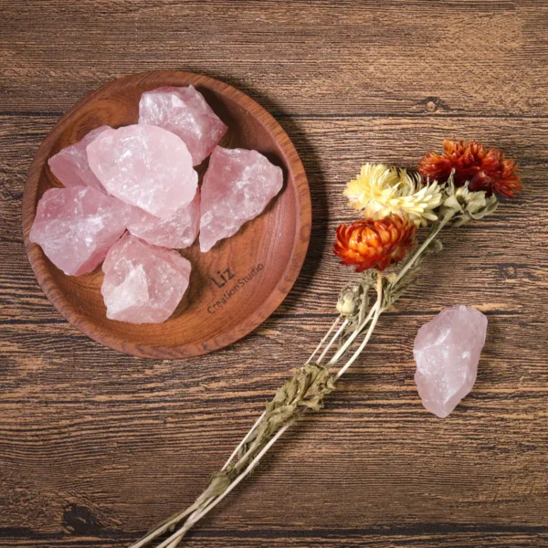 May include: A collection of raw, pink rose quartz crystals displayed in a wooden bowl and scattered on a wooden surface. Dried flowers in shades of orange and May include: A collection of raw, pink rose quartz crystals displayed in a wooden bowl and scattered on a wooden surface. Dried flowers in shades of orange and yellow are also present, alongside a single rose quartz piece.