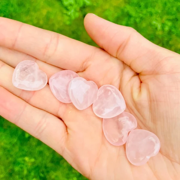 May include: A hand holding several heart-shaped rose quartz stones. The stones are a soft pink color and appear smooth and polished. The background is a blurre May include: A hand holding several heart-shaped rose quartz stones. The stones are a soft pink color and appear smooth and polished. The background is a blurred green.