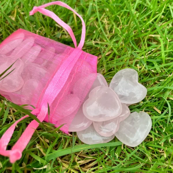 May include: A pink mesh drawstring bag spilling heart-shaped rose quartz stones onto green grass. The stones are a pale pink color and appear smooth and polish May include: A pink mesh drawstring bag spilling heart-shaped rose quartz stones onto green grass. The stones are a pale pink color and appear smooth and polished. The bag is a vibrant pink.